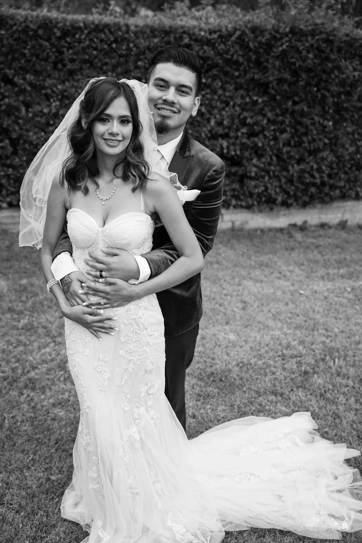 Black and white portrait of a groom embracing his bride from behind, her long train spread across the lawn